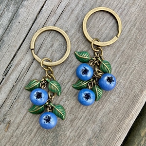 May include: Two keyrings featuring clusters of round, light blue berries with dark centres and textured green leaves. Each keyring is attached to a circular, antique brass-toned metal ring by a short chain, showcasing a botanical design.