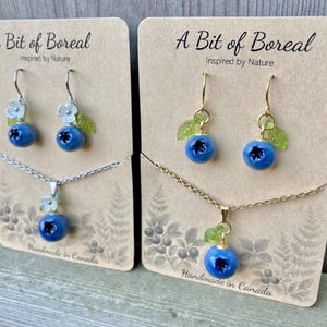 May include: Two sets of jewellery on cardboard displays labelled A Bit of Boreal. Each set includes a necklace and earrings featuring blue berry charms with small white flowers and green leaves. One set has silver-toned metal, the other gold-toned.