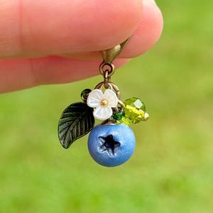 May include: A charm featuring a matte blue blueberry with a star-shaped indentation, a small white flower with a gold centre, a dark green metal leaf, and a faceted lime green bead, all attached to a bronze-toned metal lobster clasp.