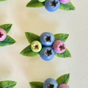 May include: A collection of small, handcrafted berry clusters made from polymer clay. Each cluster features two blue berries, one light green berry, and one purple berry, all nestled among several detailed green leaves with visible veins.