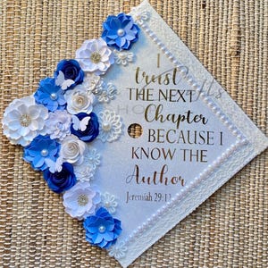 May include: A square graduation cap topper with a white glitter surface, decorated with blue and white paper flowers, small white butterflies, and pearl accents. The text reads: I trust THE NEXT Chapter BECAUSE I KNOW THE Author Jeremiah 29:11.