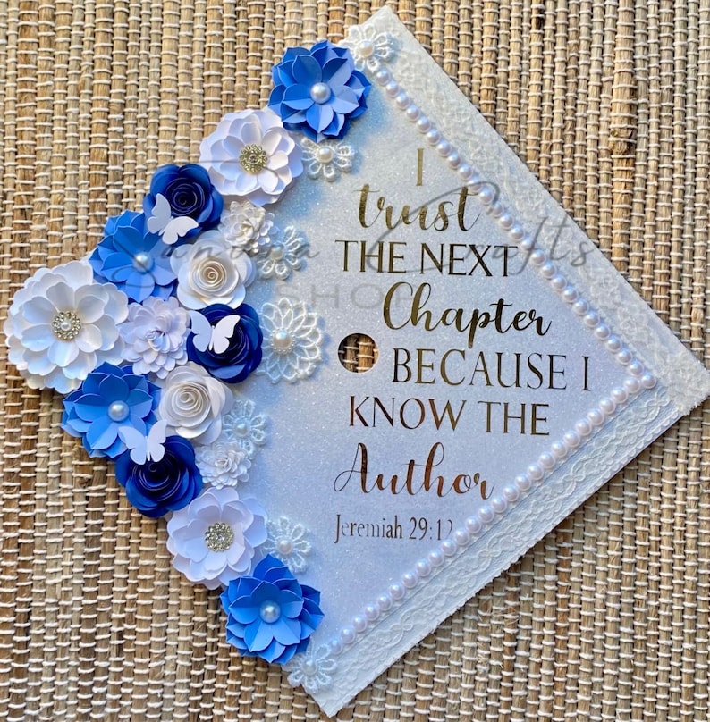 May include: A square graduation cap topper with a white glitter surface, decorated with blue and white paper flowers, small white butterflies, and pearl accents. The text reads: I trust THE NEXT Chapter BECAUSE I KNOW THE Author Jeremiah 29:11.