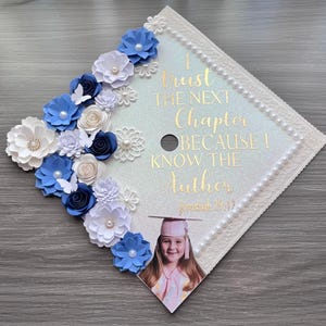 May include: A square graduation cap topper featuring a glittery white surface decorated with blue and white paper flowers, pearls, and butterflies. Gold text reads: I Trust THE NEXT Chapter BECAUSE I KNOW THE Author Jeremiah 29:11, with a photo of a graduate.