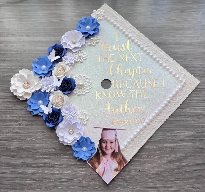 May include: A square graduation cap topper featuring a glittery white surface decorated with blue and white paper flowers, pearls, and butterflies. Gold text reads: I Trust THE NEXT Chapter BECAUSE I KNOW THE Author Jeremiah 29:11, with a photo of a graduate.