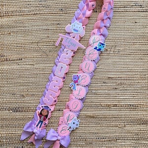 May include: A graduation lei in shades of pink and purple, adorned with character images and the text "Class of 2023". The lei features decorative bows and is set against a woven background.