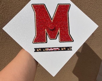 May include: A white diamond-shaped graduation cap topper featuring a large letter M filled with red rhinestones and outlined in gold. Below the letter is a horizontal black banner with a small, multi-colored graphic pattern of a flag.