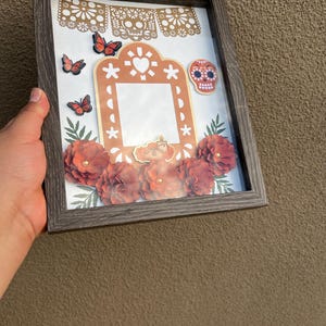 May include: A shadow box frame featuring a central rectangular cutout, surrounded by paper cutouts of monarch butterflies, a sugar skull, a candle, and dark orange paper flowers. A decorative banner with skull motifs hangs at the top.