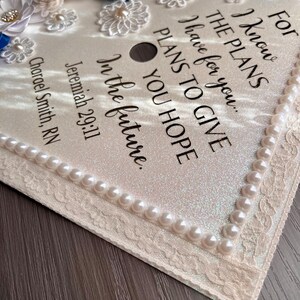 May include: A graduation cap topper featuring a shimmering cream surface decorated with blue and cream fabric flowers, pearl accents, and lace trim. The text reads: For I know THE PLANS I have for you... PLANS TO GIVE YOU HOPE In the future. Jeremiah 29:11 Charael Smith, RN.