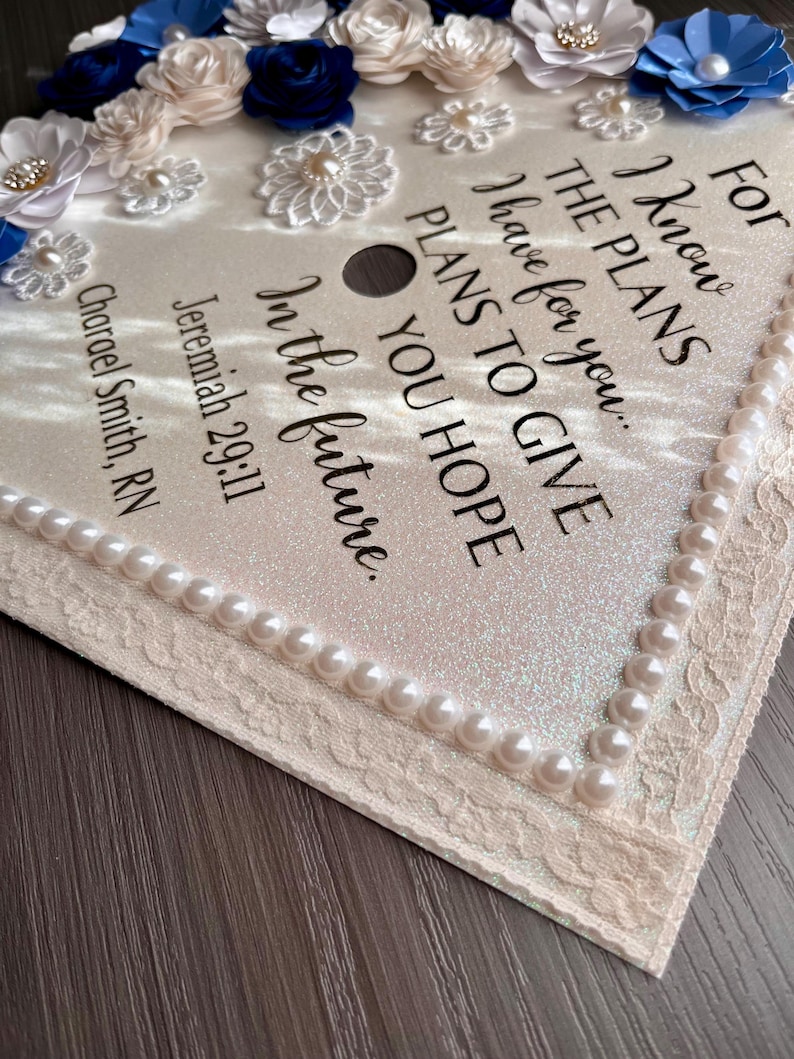 May include: A graduation cap topper featuring a shimmering cream surface decorated with blue and cream fabric flowers, pearl accents, and lace trim. The text reads: For I know THE PLANS I have for you... PLANS TO GIVE YOU HOPE In the future. Jeremiah 29:11 Charael Smith, RN.