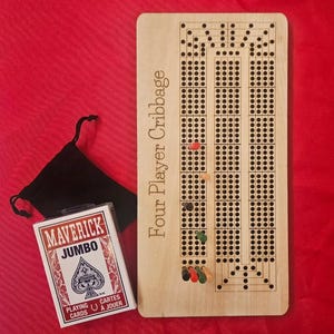 Op de afbeelding: Een houten Four Player Cribbage-bord met een pak Maverick Jumbo speelkaarten en een zwart zakje met trekkoord. Het cribbagebord heeft meerdere rijen gaten om te pinnen en is gemaakt van lichtgekleurd hout. De speelkaarten zitten in een wit en rood doosje.