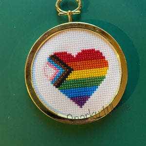 Puede incluir: Un aro de bordado dorado con un fondo de tela blanca que presenta un corazón arcoíris con una franja marrón, azul, rosa y blanca en el lado izquierdo.