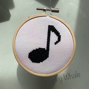 May include: A white cross-stitch embroidery hoop with a black musical note design. The hoop is made of wood and has a metal stand.