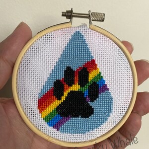 Op de afbeelding: Een regenboog-pootafdruk-kruissteekontwerp in een houten borduurring. De poot is zwart en de regenboog is rood, oranje, geel, groen, blauw, indigo en violet. De achtergrond is lichtblauw.
