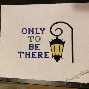 May include: Cross-stitch embroidery on white fabric with the text "ONLY TO BE THERE" in colourful letters and a black silhouette of a street lamp with a yellow light.