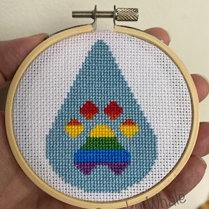 Op de afbeelding: Een kruissteekontwerp met een regenboogpootafdruk in een houten borduurring. De potafdruk is blauw met een regenboogvormige boog en rode pootkussentjes.