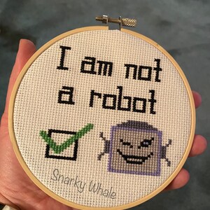 May include: A white embroidery hoop with the text "I am not a robot" in black. A green checkmark is next to a purple robot face with a winking eye. The text "Sharky Whale" is at the bottom of the hoop.