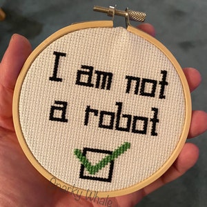 May include: A white embroidery hoop with the text "I am not a robot" and a green checkmark in a black square. The text "Snarky Whale" is written below the checkmark.