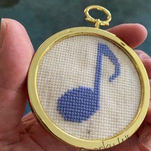 May include: A small, round, gold-framed embroidery hoop with a light blue musical note stitched on a white linen background.