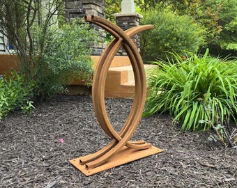 Puede incluir: Una escultura de metal abstracta y desgastada con un acabado marrón óxido. La escultura presenta líneas curvas que se cruzan y una base rectangular, situada al aire libre en un jardín. Una pieza decorativa para espacios exteriores.
