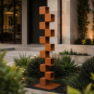 May include: A tall, vertical outdoor sculpture composed of ten stacked, open-faced metal cubes with a rusted, textured finish. The cubes are arranged in an alternating, offset pattern, supported by a flat, square metal base on the ground.
