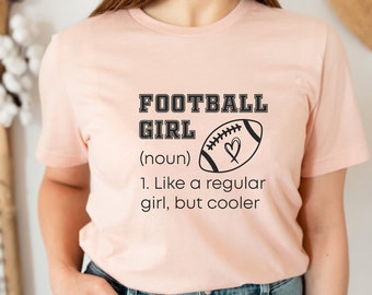May include: A light pink t-shirt with the text "FOOTBALL GIRL" above a football graphic with a heart. Below the graphic is the definition: "1. Like a regular girl, but cooler."