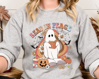 Puede incluir: Una sudadera gris claro con un gráfico colorido que dice "READ IN PEACE". El gráfico presenta un fantasma leyendo un libro, rodeado de calabazas, flores, murciélagos y un arcoíris. El diseño tiene un estilo retro y vintage.