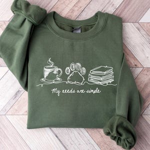 May include: An olive green sweatshirt with a white graphic of a coffee cup, paw print, and books. The text "My needs are simple" is below the graphic. The sweatshirt is folded on a light wood surface.