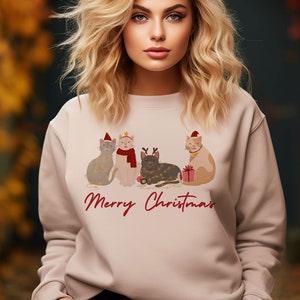 以下が含まれることがあります： 4匹の猫がクリスマスの飾りを身につけたデザインの、明るいベージュ色のスウェットシャツ。「Merry Christmas」の文字が赤字で書かれています。猫たちは帽子やスカーフ、ライトで飾られています。