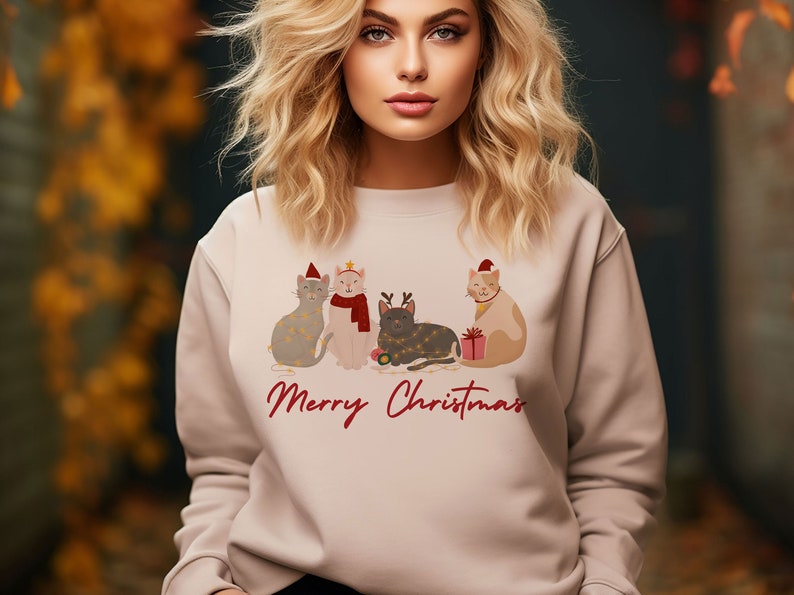 以下が含まれることがあります： 4匹の猫がクリスマスの飾りを身につけたデザインの、明るいベージュ色のスウェットシャツ。「Merry Christmas」の文字が赤字で書かれています。猫たちは帽子やスカーフ、ライトで飾られています。