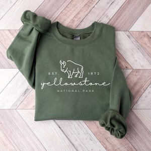 May include: Dark green sweatshirt featuring a white bison graphic and text that reads "Yellowstone National Park". The text "Est. 1872" is above the name. The garment is displayed on a light wood-effect surface.