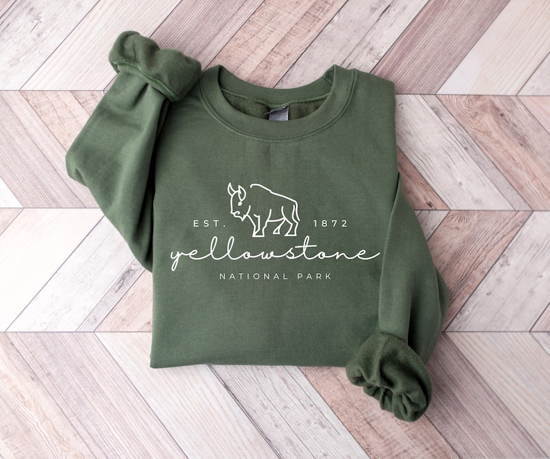 May include: Dark green sweatshirt featuring a white bison graphic and text that reads "Yellowstone National Park". The text "Est. 1872" is above the name. The garment is displayed on a light wood-effect surface.