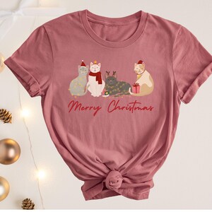 以下が含まれることがあります： クリスマスアクセサリーを身に着けた4匹の猫のグラフィックが特徴の、くすんだピンク色のTシャツ。「Merry Christmas」の文字が筆記体で書かれています。