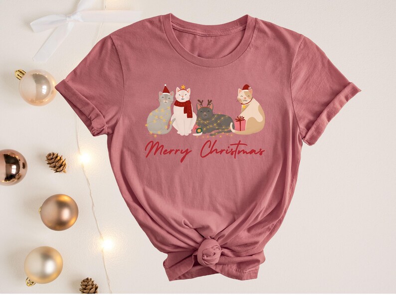 以下が含まれることがあります： クリスマスアクセサリーを身に着けた4匹の猫のグラフィックが特徴の、くすんだピンク色のTシャツ。「Merry Christmas」の文字が筆記体で書かれています。