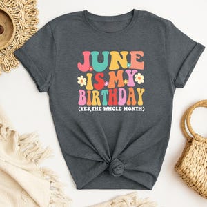 May include: A dark gray t-shirt with the text "June is my birthday (yes, the whole month)" in colorful retro lettering with flowers.