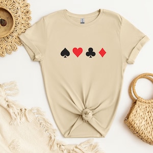 Op de afbeelding: Een beige t-shirt met de vier speelkaartsymbolen: schoppen, harten, klaveren en ruiten. De symbolen zijn zwart en rood. Het shirt is gemaakt van een zacht materiaal en heeft opgerolde mouwen.