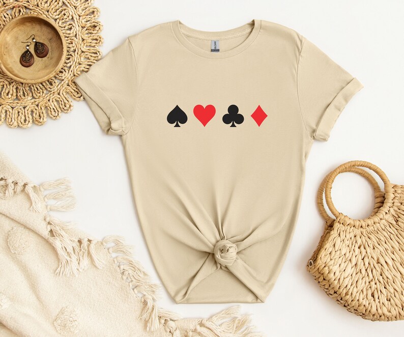 Op de afbeelding: Een beige t-shirt met de vier speelkaartsymbolen: schoppen, harten, klaveren en ruiten. De symbolen zijn zwart en rood. Het shirt is gemaakt van een zacht materiaal en heeft opgerolde mouwen.
