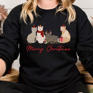 以下が含まれることがあります： クリスマスのデザインが施された黒いスウェットシャツ。サンタ帽や電飾など、お祝いのアクセサリーを身につけた4匹の猫が描かれています。「Merry Christmas」の文字が赤色の筆記体で下に書かれています。