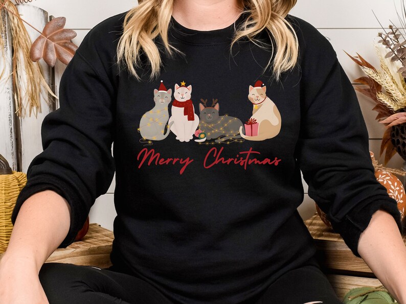 以下が含まれることがあります： クリスマスのデザインが施された黒いスウェットシャツ。サンタ帽や電飾など、お祝いのアクセサリーを身につけた4匹の猫が描かれています。「Merry Christmas」の文字が赤色の筆記体で下に書かれています。