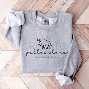 May include: A grey sweatshirt featuring the words "Yellowstone National Park" in a script font, with a bison graphic. The text "Est. 1872" is above the word "Yellowstone". The sleeves are rolled up.