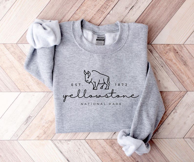 May include: A grey sweatshirt featuring the words "Yellowstone National Park" in a script font, with a bison graphic. The text "Est. 1872" is above the word "Yellowstone". The sleeves are rolled up.