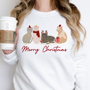 以下が含まれることがあります： クリスマスのデザインが施された白いスウェットシャツ。サンタ帽やフェアリーライトなど、お祝いのアクセサリーを身に着けた4匹の漫画の猫が特徴です。「Merry Christmas」の文字が猫の下に赤いスクリプトで書かれています。温かい飲み物が人の手に持たれています。