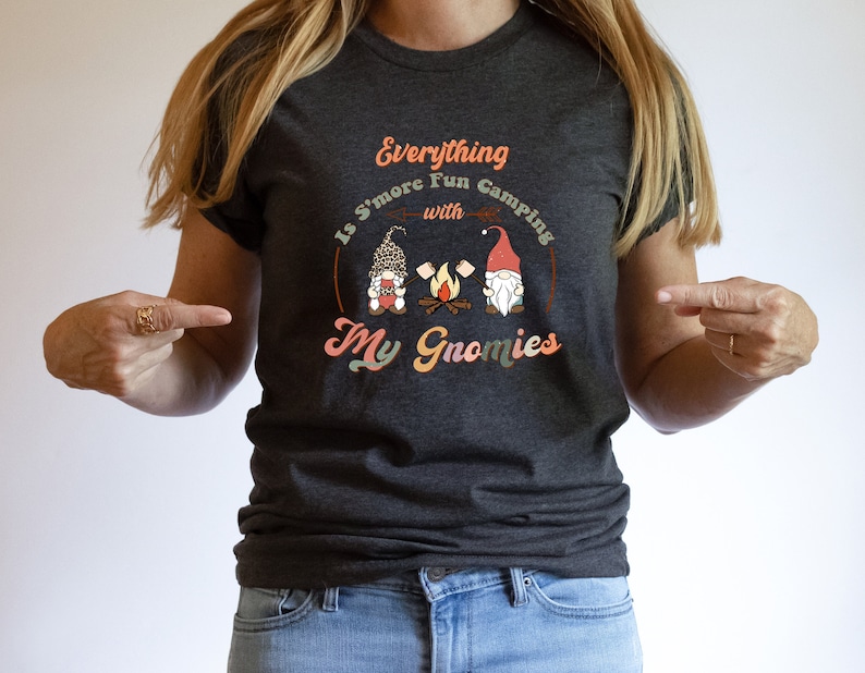 May include: A dark gray t-shirt with the text "Everything is S'more Fun Camping with My Gnomies" in a retro font. The text is surrounded by a circle with a campfire and two gnomes.