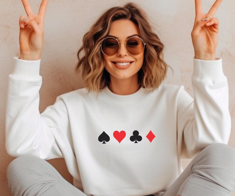 Op de afbeelding: Witte sweatshirt met speelkaartsymbolen: schoppen, harten, klaveren en ruiten. De symbolen zijn zwart en rood. De persoon op de foto draagt een zonnebril en maakt een vredesteken.