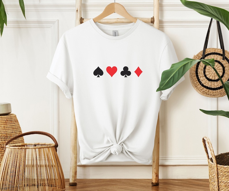 Op de afbeelding: Wit T-shirt met een ontwerp van speelkaartsymbolen: een zwarte schoppen, een rode harten, een zwarte klaveren en een rode ruiten. Het shirt hangt aan een houten hanger tegen een neutrale achtergrond.