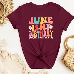 May include: A maroon t-shirt with the text "June is my birthday (yes, the whole month)" in colorful retro lettering.