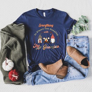 May include: A navy blue t-shirt with a graphic of three gnomes around a campfire. The text on the shirt reads "Everything is S'more Fun Camping with My Gnomies".