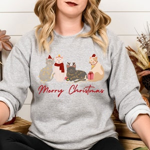 以下が含まれることがあります： クリスマスをテーマにしたデザインのライトグレーのスウェットシャツ。サンタ帽やフェアリーライトなどのお祝いのアクセサリーを身に着けた4匹の漫画の猫が特徴です。「Merry Christmas」の文字が赤色のスクリプトで猫の下に書かれています。