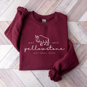 May include: A maroon sweatshirt featuring the text "Yellowstone National Park" and a white bison graphic. The sweatshirt has a crew neck and long sleeves. The text "EST. 1872" is above the word "Yellowstone". It rests on a light wood surface.