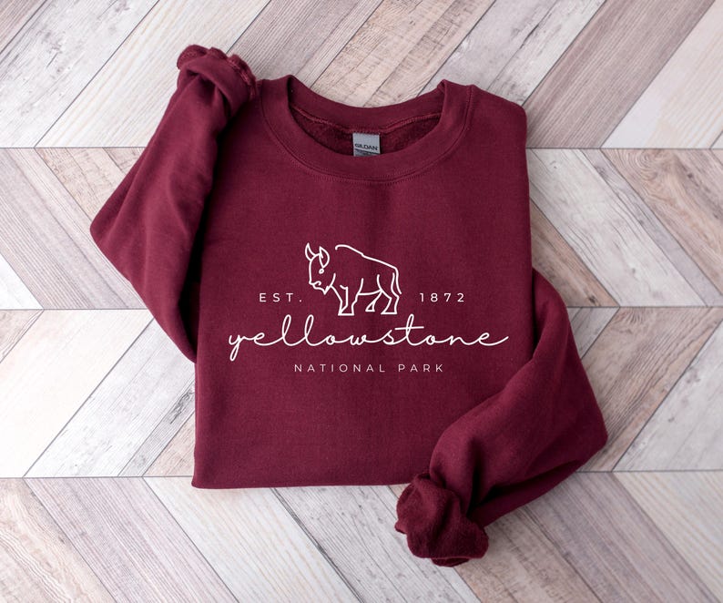May include: A maroon sweatshirt featuring the text "Yellowstone National Park" and a white bison graphic. The sweatshirt has a crew neck and long sleeves. The text "EST. 1872" is above the word "Yellowstone". It rests on a light wood surface.