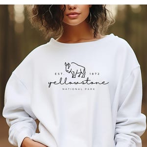 May include: White sweatshirt featuring a black bison graphic and the text "Yellowstone National Park" and "Est. 1872". The top has a crew neck and long sleeves. The design is centrally placed on the chest.